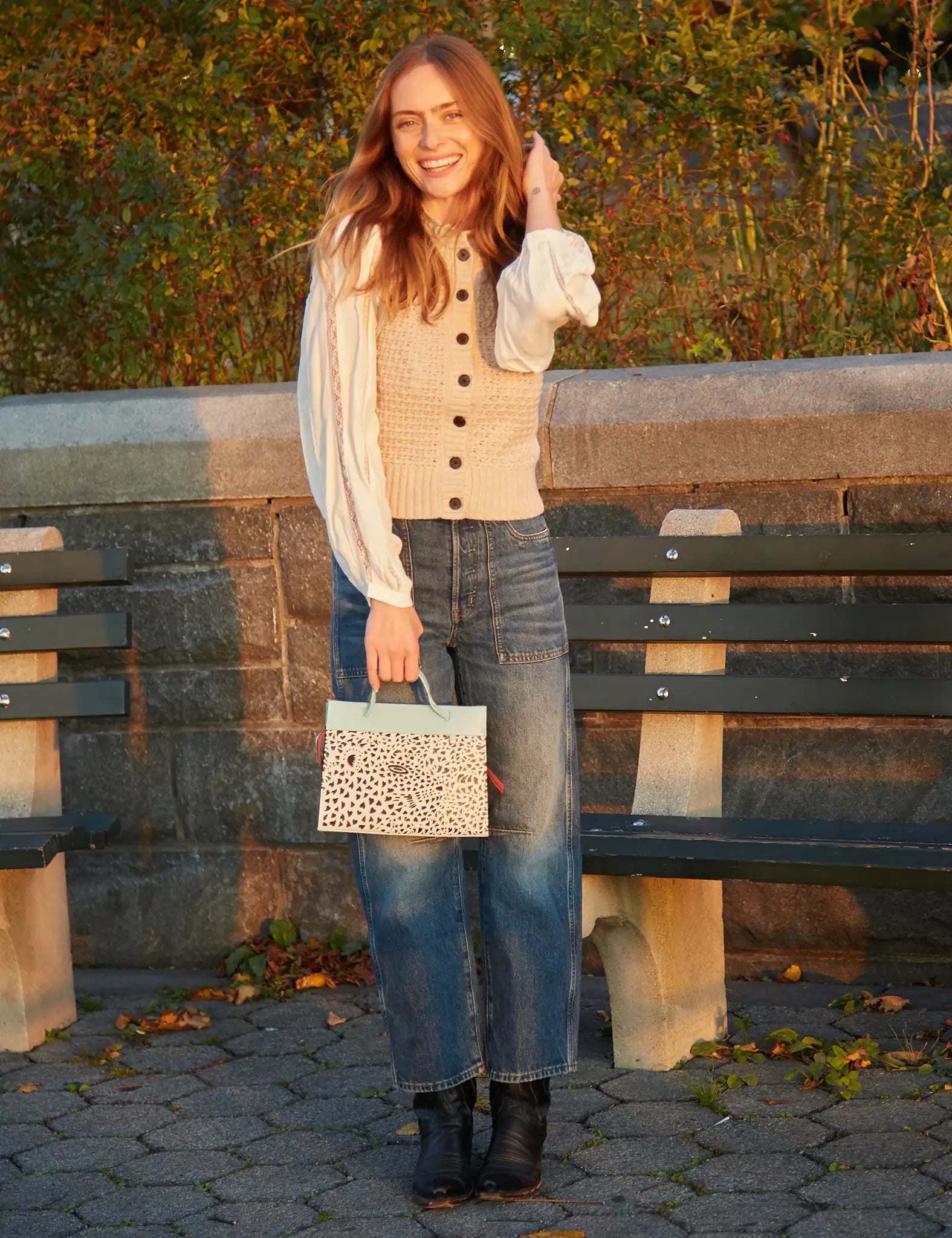 Woman standing outdoors holding a patterned handbag, wearing a beige cardigan and blue jeans.