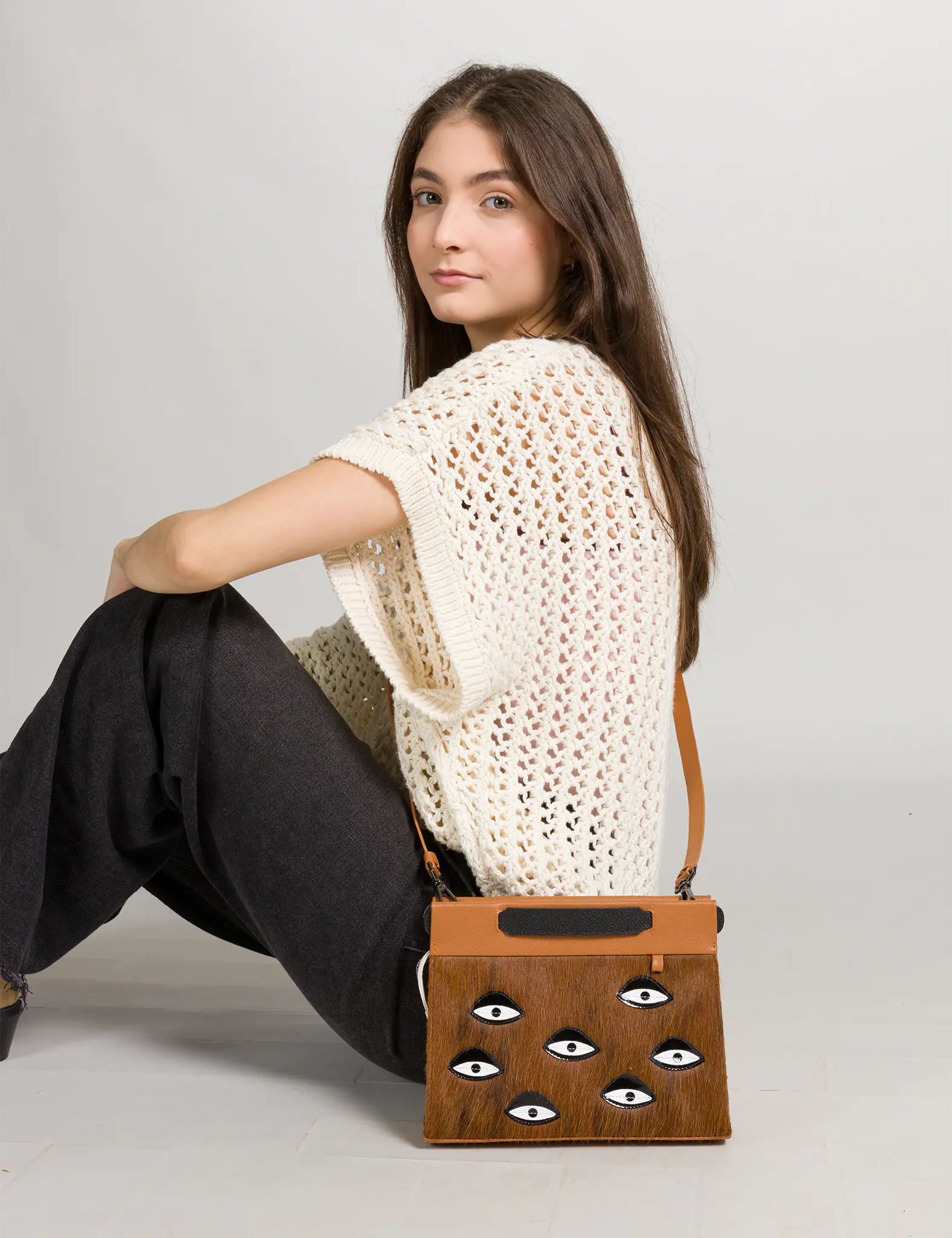 Woman sitting with a fur brown handbag featuring eye patterns on a plain background