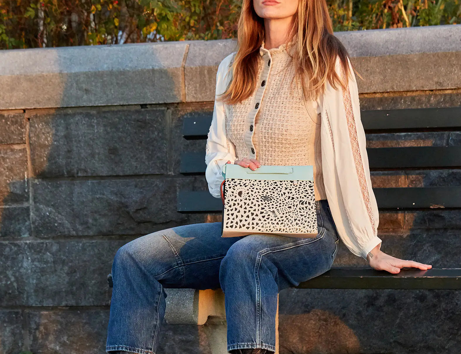 A woman sitting on a park bench during golden hour, holding a beige and light blue clutch with a black speckled pattern, wearing jeans, boots, and a cream blouse.