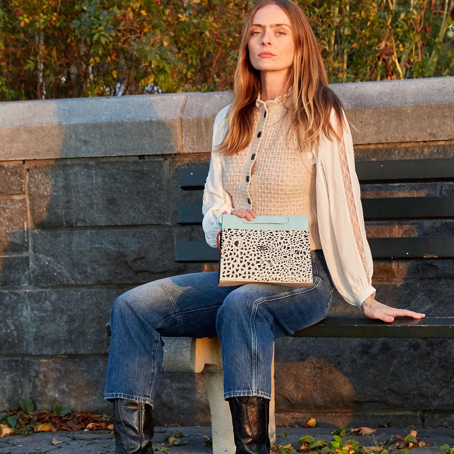 A woman sitting on a park bench during golden hour, holding a beige and light blue clutch with a black speckled pattern, wearing jeans, boots, and a cream blouse.
