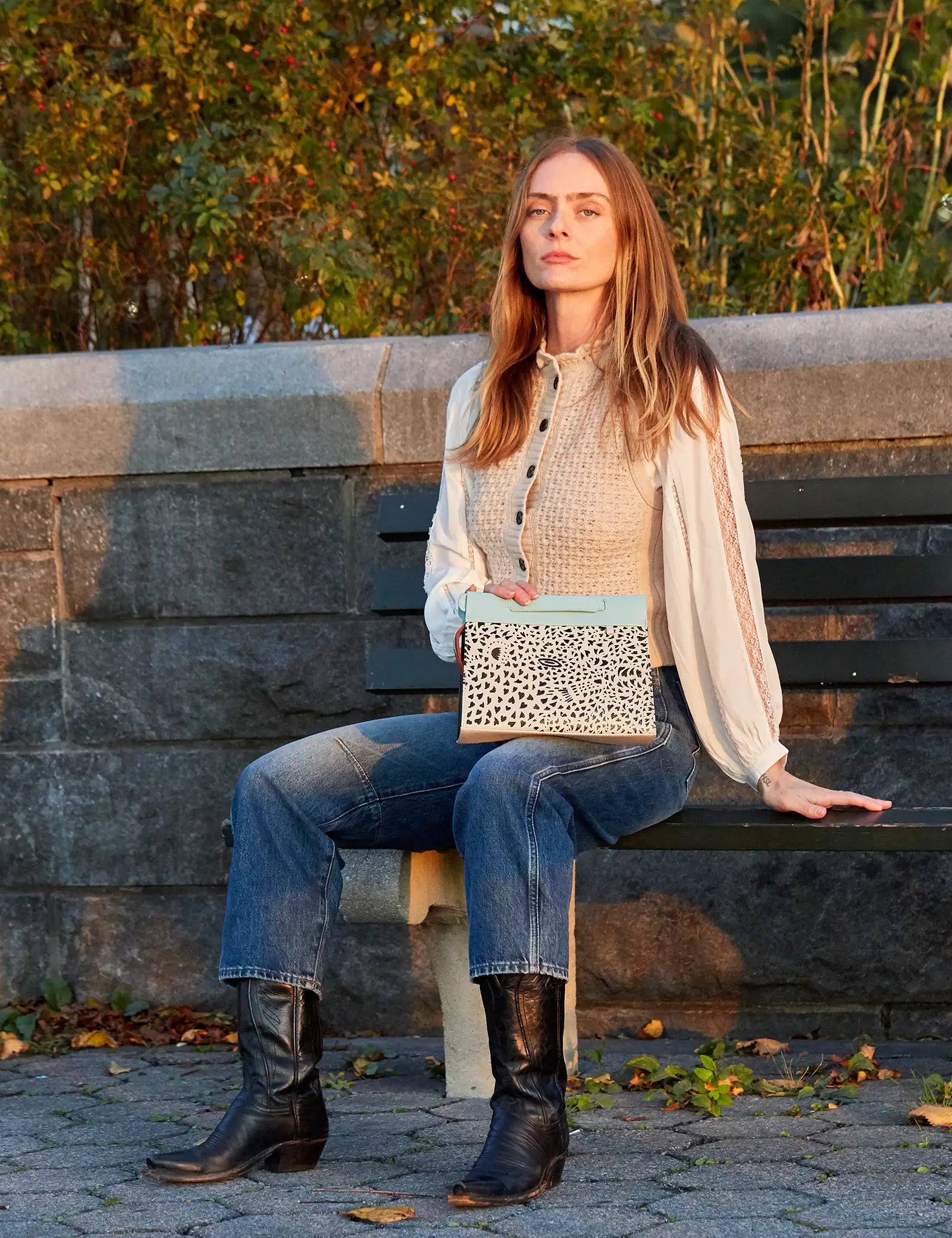 A woman sitting on a park bench during golden hour, holding a beige and light blue clutch with a black speckled pattern, wearing jeans, boots, and a cream blouse.
