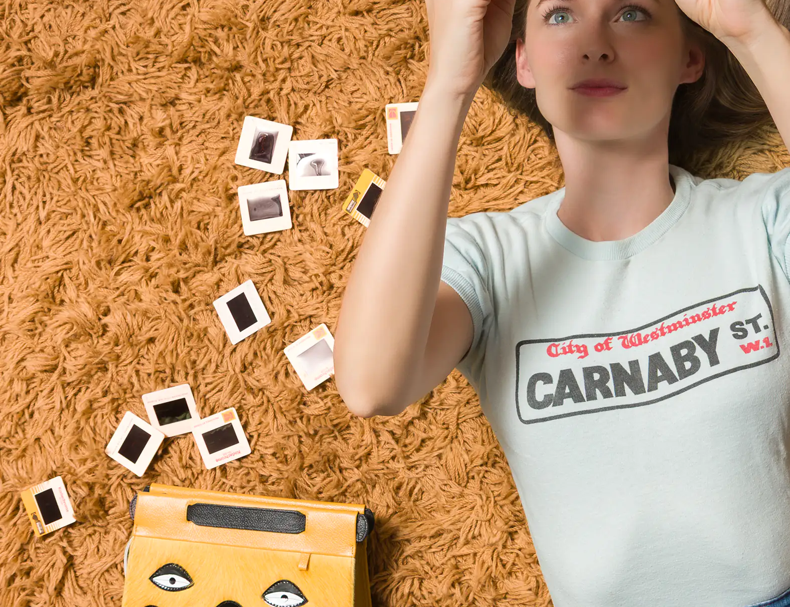 A woman lying on a shaggy orange carpet, holding up photo slides while a yellow handbag with eye motifs rests beside her.