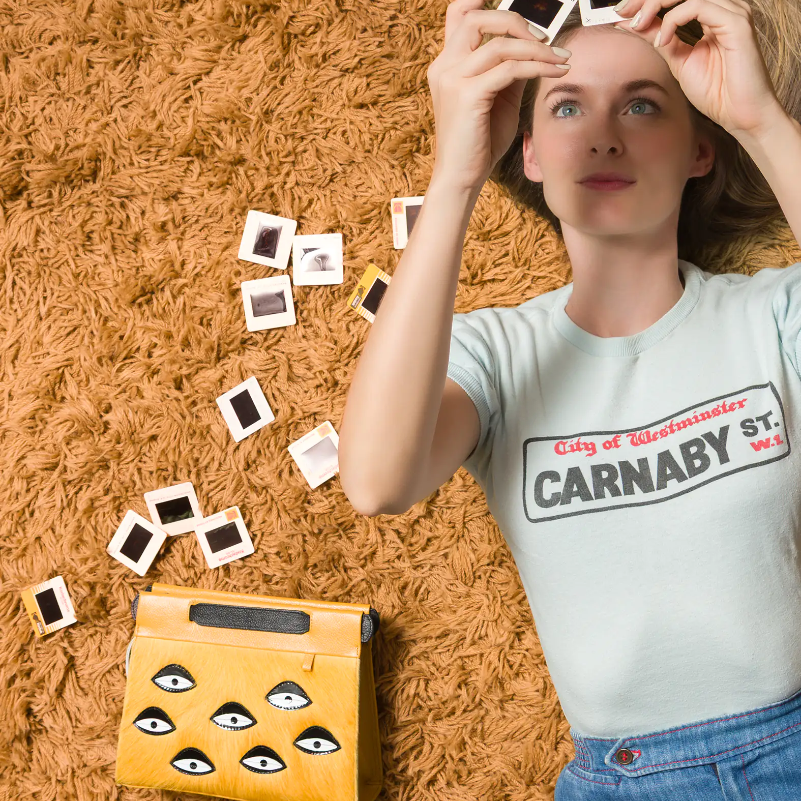 A woman lying on a shaggy orange carpet, holding up photo slides while a yellow handbag with eye motifs rests beside her.