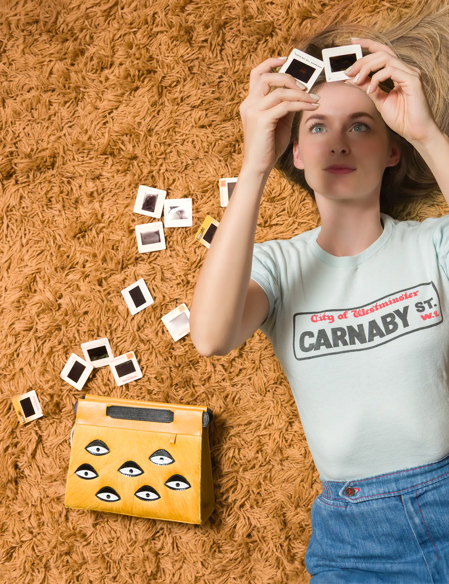 A woman lying on a shaggy orange carpet, holding up photo slides while a yellow handbag with eye motifs rests beside her.