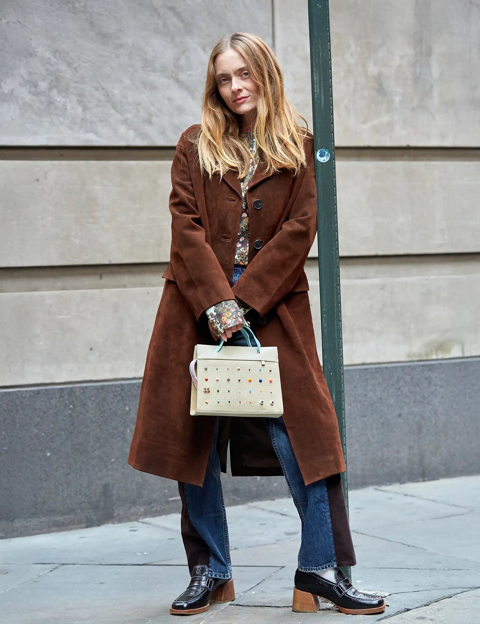 A woman wearing a long brown coat, floral blouse, and two-tone jeans stands on a city sidewalk holding a beige handbag adorned with colorful studs, exuding a casual and stylish fall look.