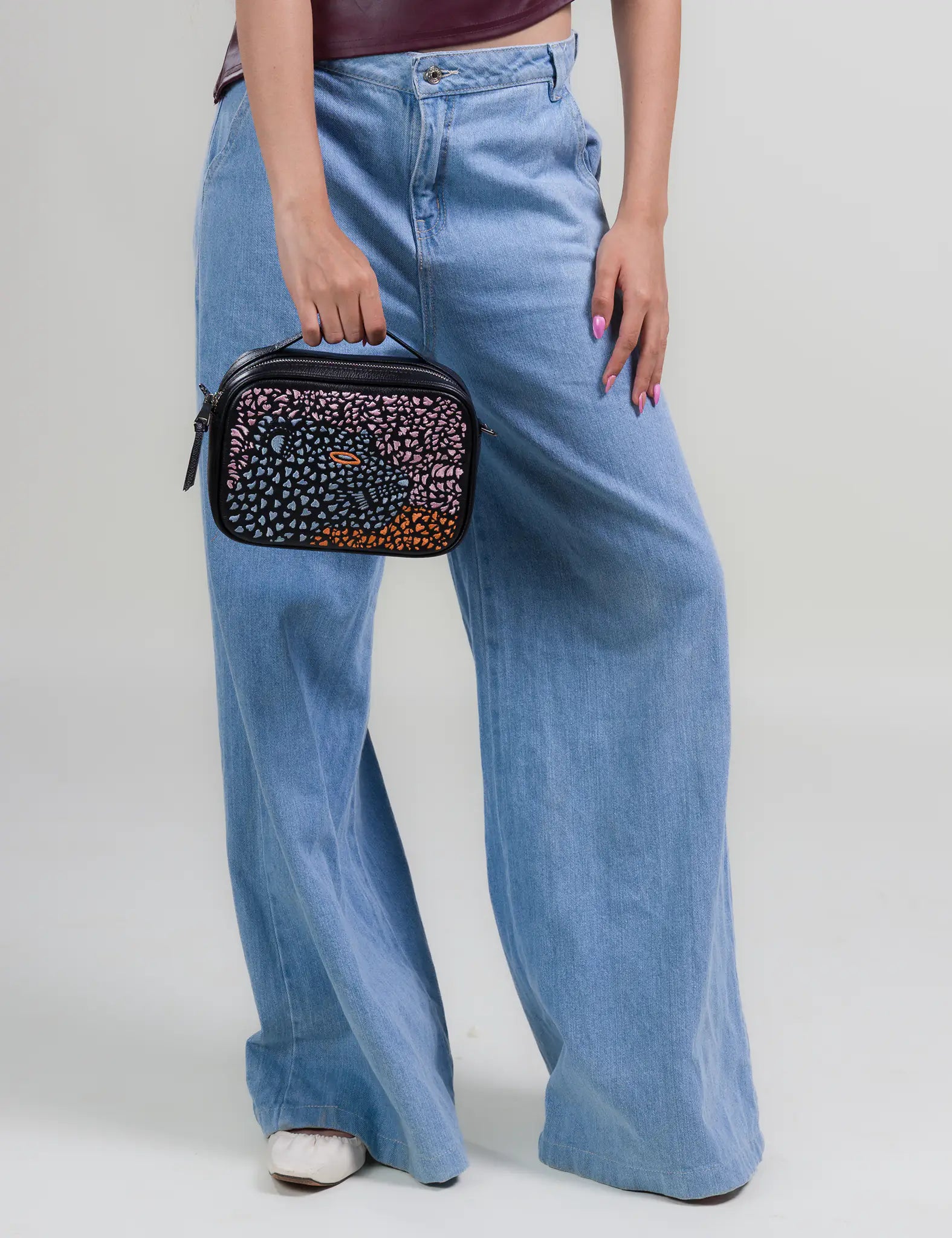 Woman in light blue wide-leg jeans holds a small black top-handle bag featuring a colorful gradient mosaic design and an eye motif, against a neutral studio background.