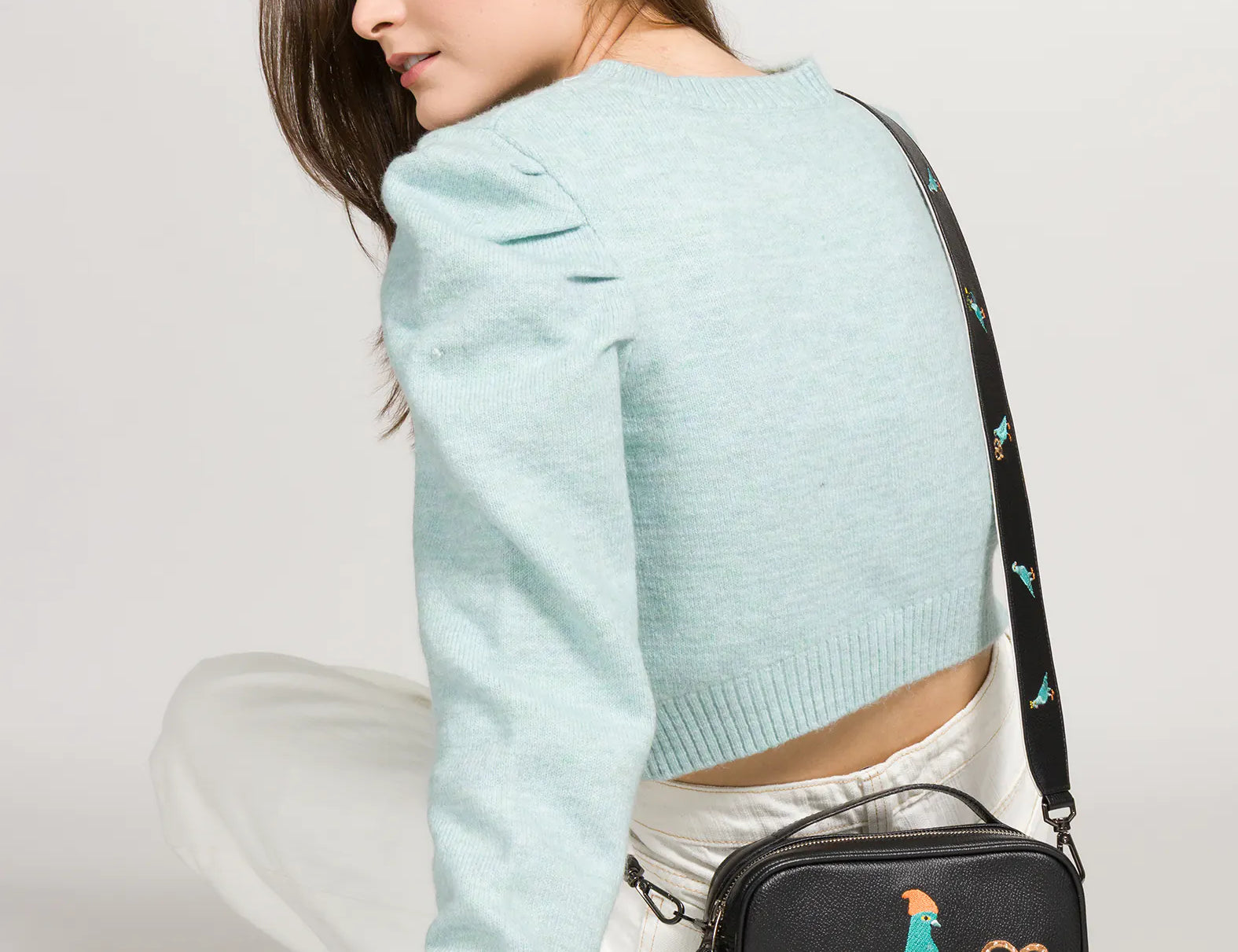Woman in a light blue cropped sweater and white pants sits turned away, looking over her shoulder while wearing a black crossbody bag with a colorful embroidered bird design.