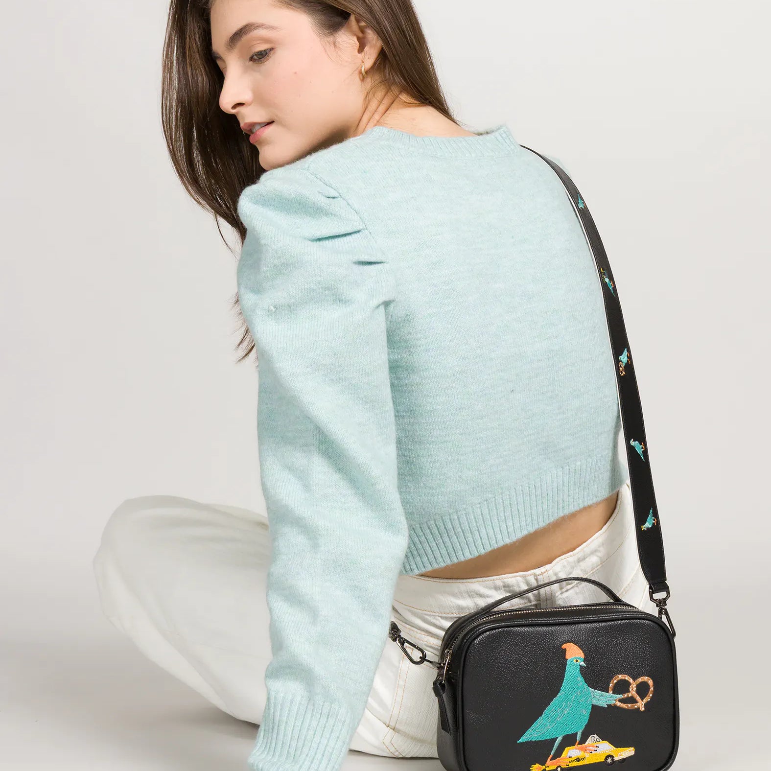 Woman in a light blue cropped sweater and white pants sits turned away, looking over her shoulder while wearing a black crossbody bag with a colorful embroidered bird design.