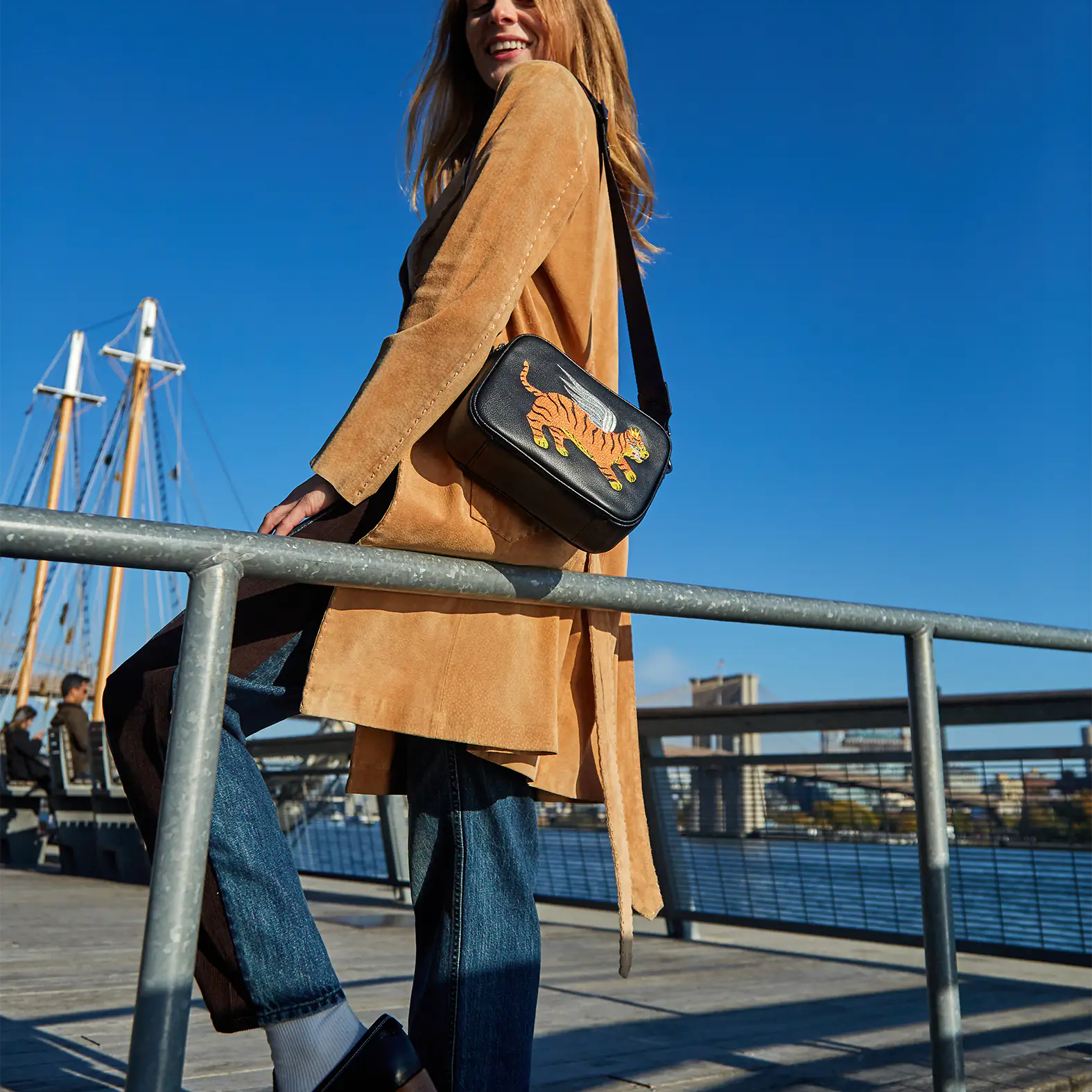 A woman standing on a waterfront boardwalk under a clear blue sky, wearing a tan coat and jeans, carrying a black crossbody bag with an embroidered tiger design.