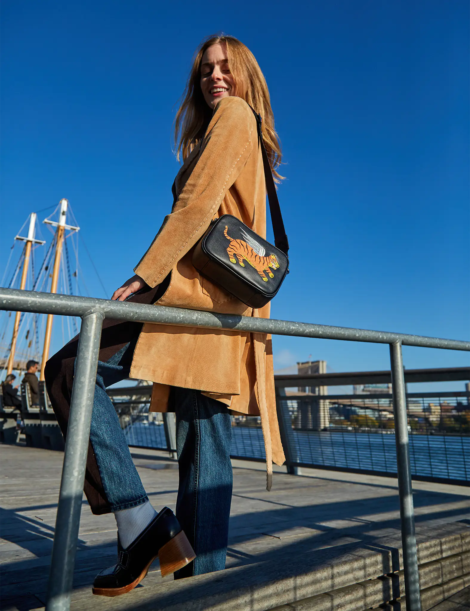 A woman standing on a waterfront boardwalk under a clear blue sky, wearing a tan coat and jeans, carrying a black crossbody bag with an embroidered tiger design.