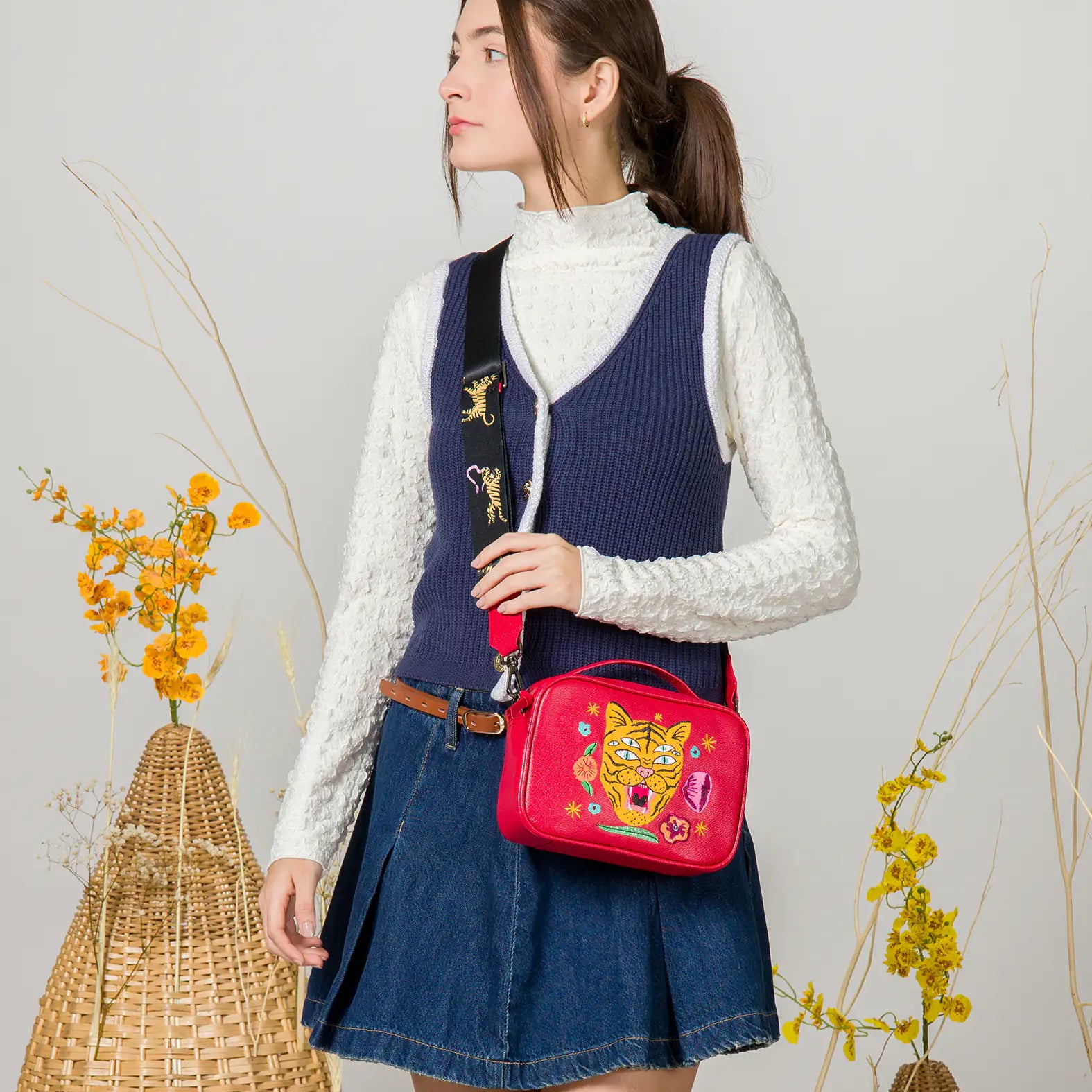 Woman in a white textured top and navy vest wears a red crossbody bag with embroidered tiger and floral design, styled with a wide black strap featuring tiger details, paired with a denim skirt.