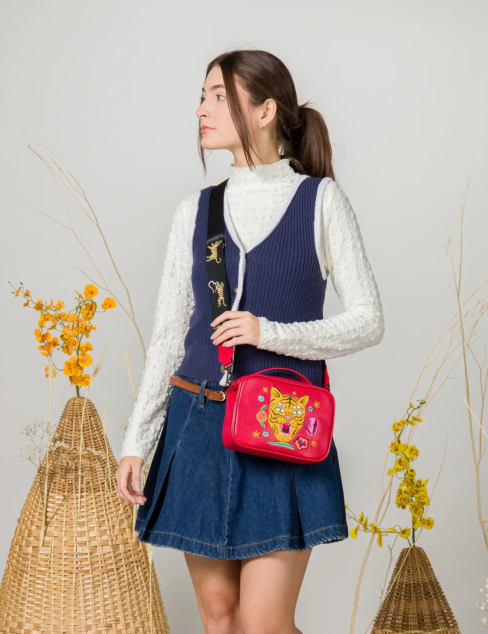 Woman in a white textured top and navy vest wears a red crossbody bag with embroidered tiger and floral design, styled with a wide black strap featuring tiger details, paired with a denim skirt.