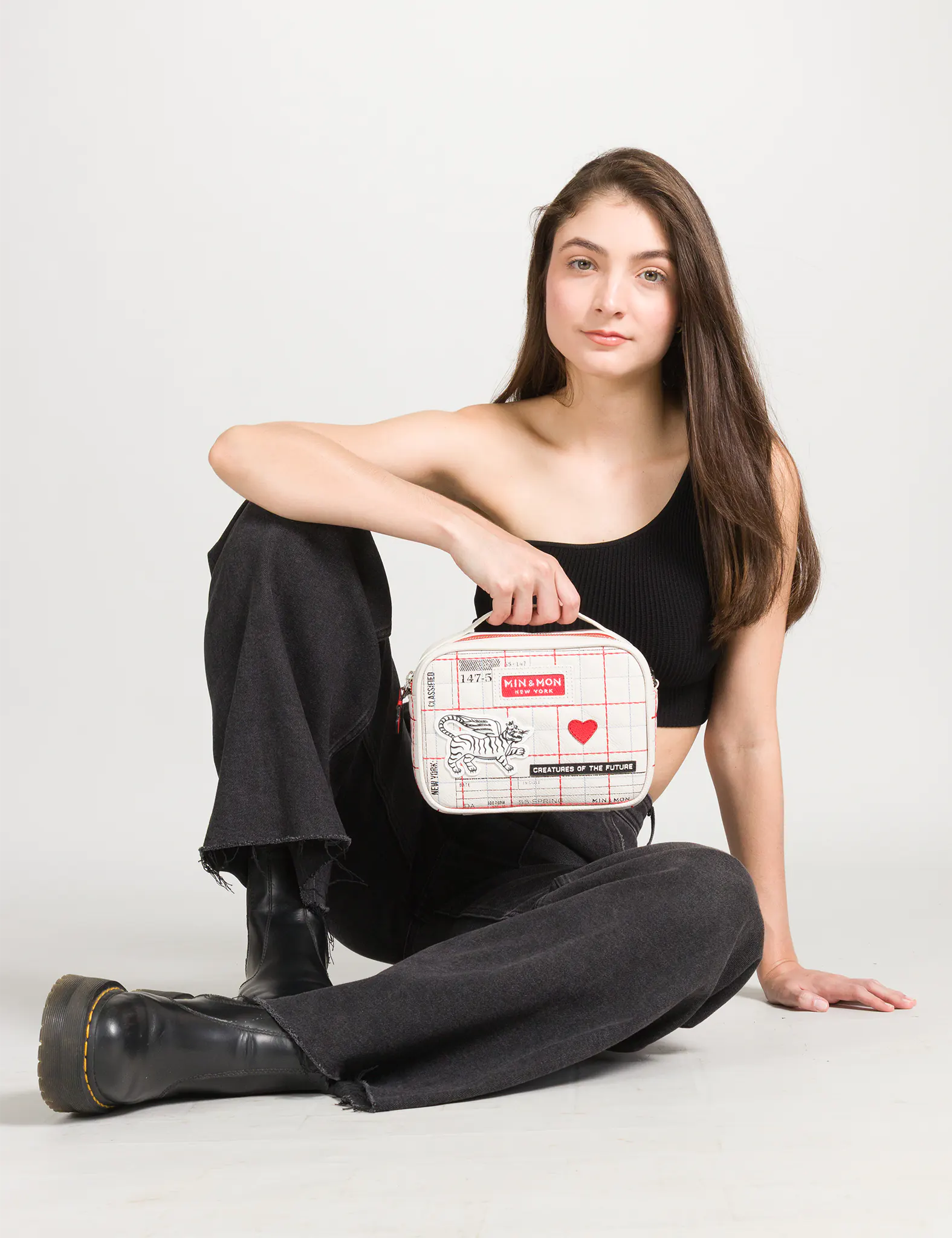 Model seated on floor in black one-shoulder top, wide-leg pants, and black boots, holding a cream mini bag with red grid print, winged tiger graphic, and red heart, against a light background.