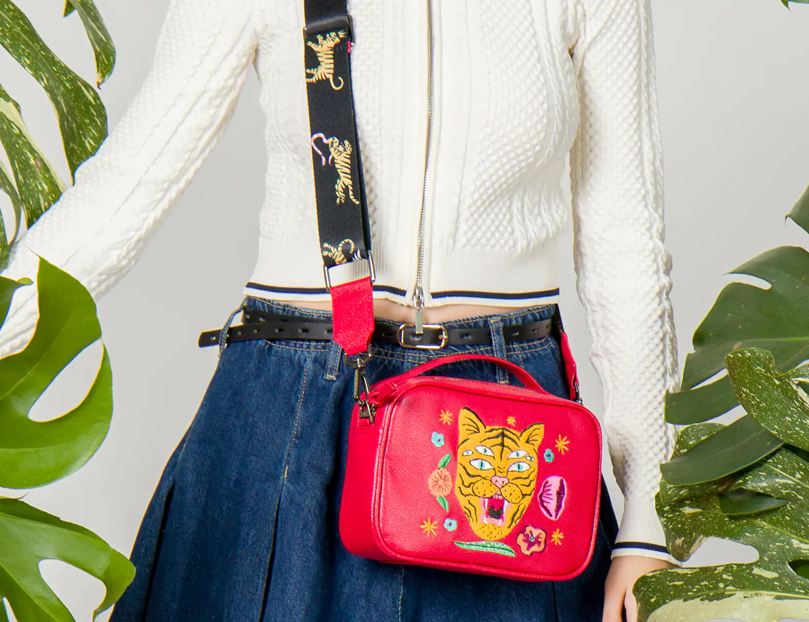 Woman in a white zip-up sweater and denim skirt wears a red crossbody bag with a bold embroidered tiger face and floral accents, styled with a wide black strap featuring tiger details.