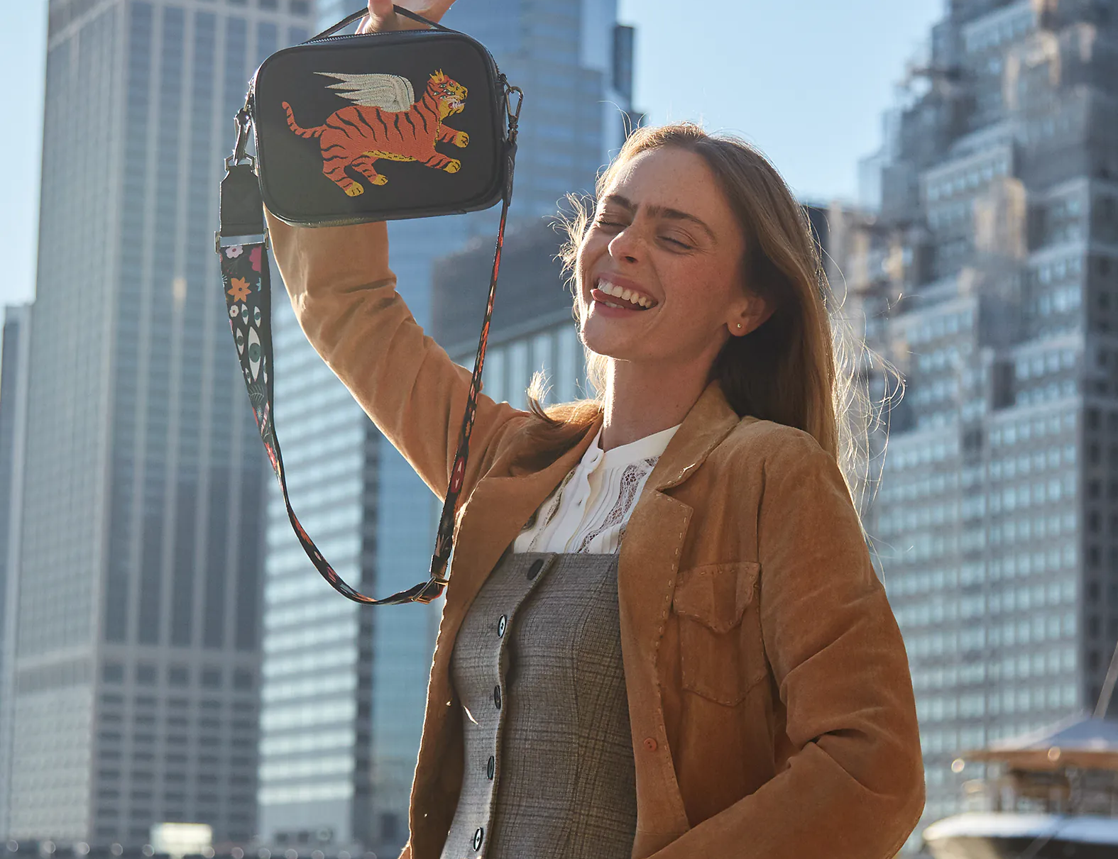 A woman smiling outdoors in a city setting, holding up a small black crossbody bag embroidered with a winged tiger design.