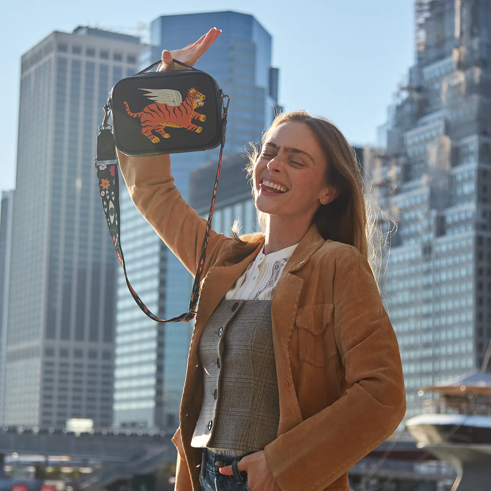 A woman smiling outdoors in a city setting, holding up a small black crossbody bag embroidered with a winged tiger design.