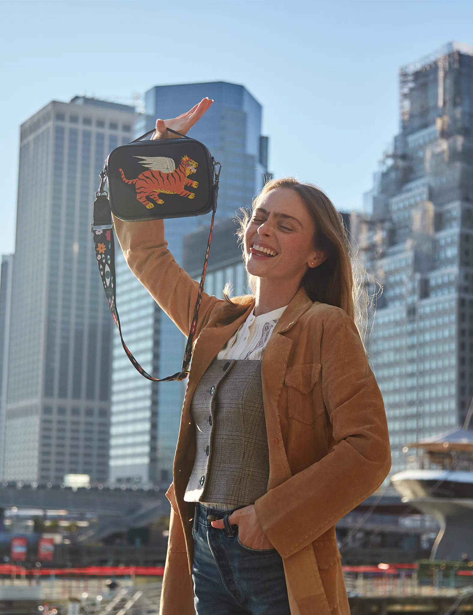 A woman smiling outdoors in a city setting, holding up a small black crossbody bag embroidered with a winged tiger design.