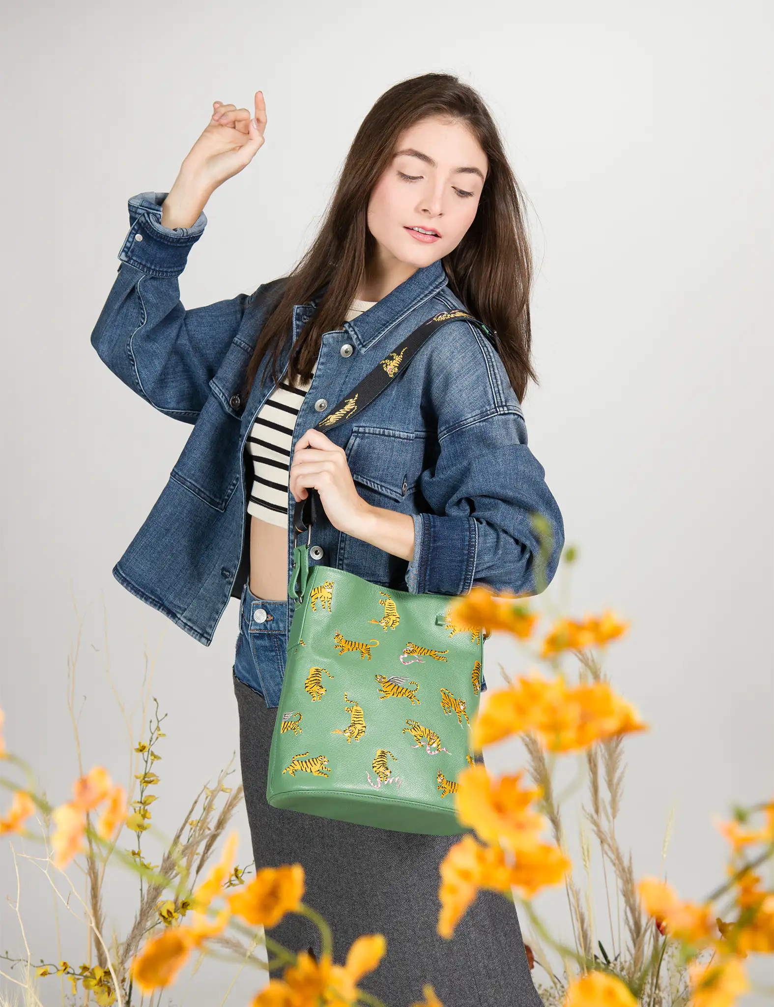 Woman posing among yellow flowers, wearing a denim jacket over a striped crop top and long skirt, carrying a green shoulder bag embroidered with golden tigers.