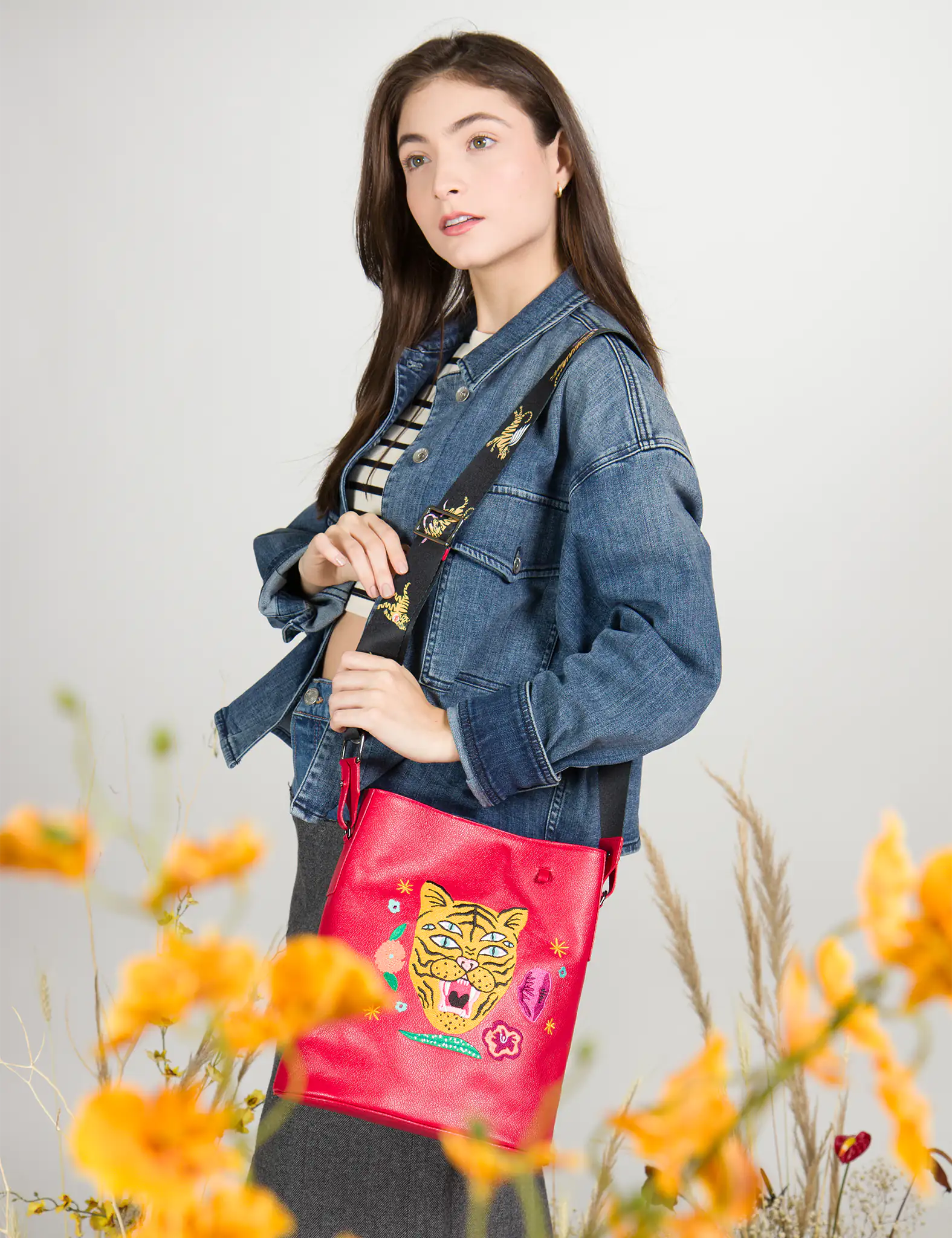 Woman in denim jacket and striped crop top carries a bold red bucket bag with a vibrant embroidered tiger face and floral motifs, paired with a wide black strap featuring golden designs.