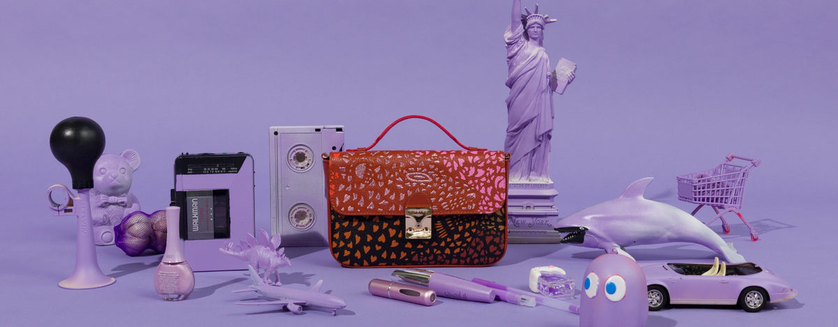 Min & Mon Amantis handbag from the Wild Heart collection, featuring a colorful animal-inspired print in orange, pink, and black, surrounded by playful purple objects and miniature New York City landmarks.
