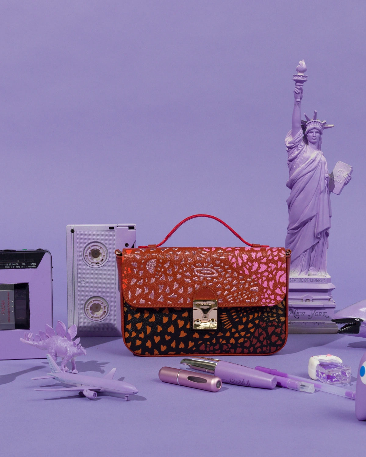 Close-up of the Min & Mon Amantis handbag from the Wild Heart collection, featuring a vibrant orange, pink, and black print with a metallic clasp, styled with purple accessories and a miniature Statue of Liberty.